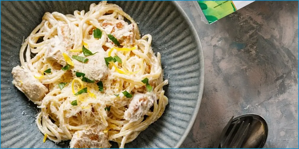 A plate of creamy gluten-free spaghetti garnished with herbs, served with a fork on a textured table.