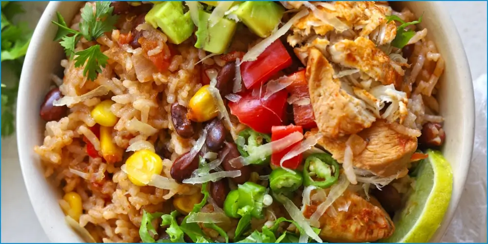 A high-protein bowl filled with rice, black beans, corn, chopped tomatoes, avocado, and grilled chicken, garnished with cilantro and a lime wedge.